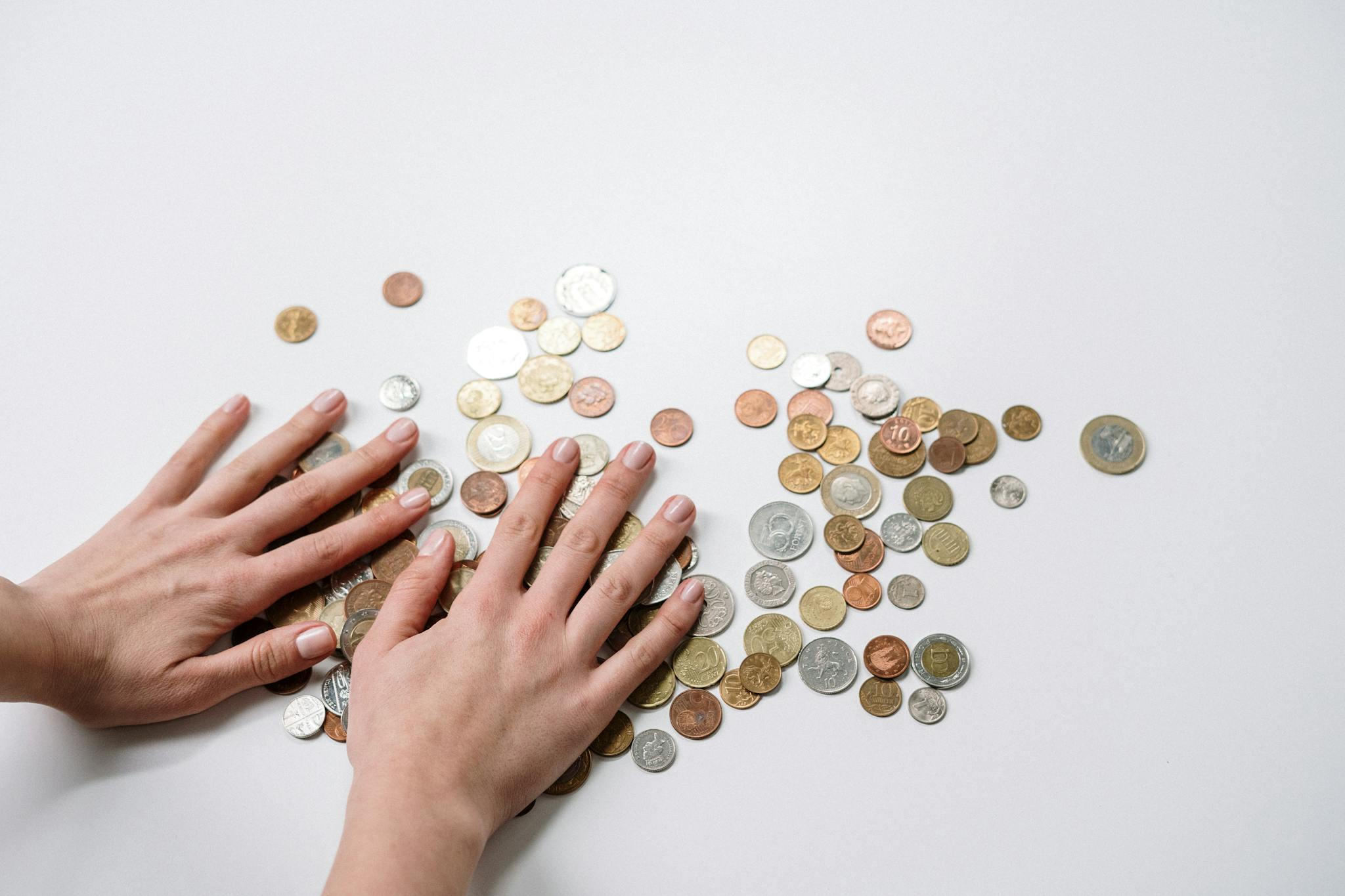Hands arranging a variety of international coins on a neutral light background.