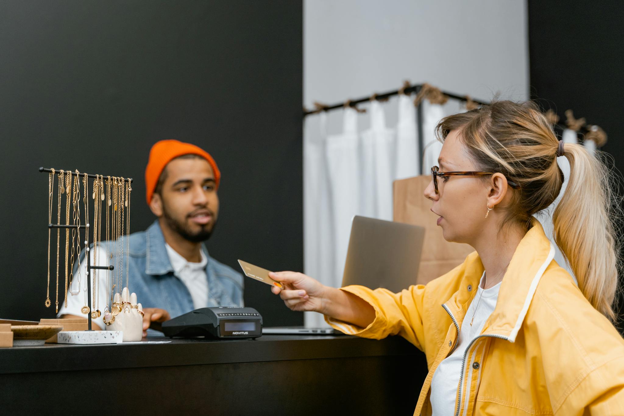 Woman in yellow jacket pays with credit card at shop counter with jewelry display.